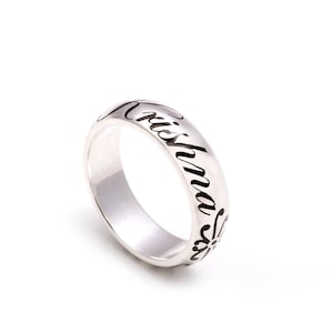 Può includere: Un anello d'argento con il nome "Cristina" inciso in corsivo. L'anello ha una superficie liscia e lucida e un motivo floreale. L'anello è posto su uno sfondo bianco.