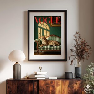 Op de afbeelding: Ingelijste kunstprint met een cheetah op een groen en wit gestreepte bank. Het rode woord "VOGUE" staat bovenaan. De achtergrond toont een groene muur, een raam en een schilderij. Stijlvolle woondecoratie.