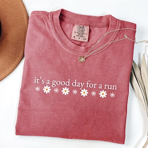 May include: A dusty rose-colored t-shirt with the phrase "it's a good day for a run" printed in white, along with daisy graphics. A silver necklace with a pendant is draped on the shirt. The shirt has a Comfort Colors tag.
