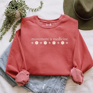 May include: A coral-colored sweatshirt with the phrase "movement is medicine" printed in white, along with white daisy graphics. The long-sleeved top is folded over a pair of blue jeans. A green hat and plant are in the background.