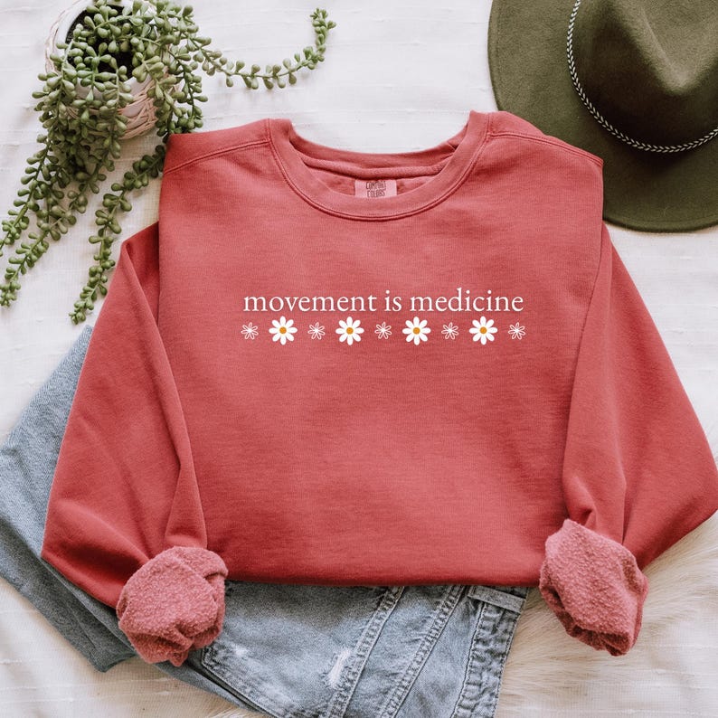 May include: A coral-colored sweatshirt with the phrase "movement is medicine" printed in white, along with white daisy graphics. The long-sleeved top is folded over a pair of blue jeans. A green hat and plant are in the background.