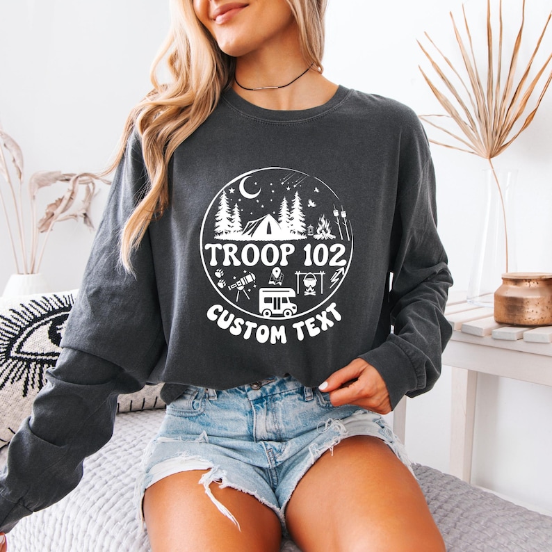 以下が含まれることがあります： テント、木、テキスト「TROOP 102 CUSTOM TEXT」を特徴とする円形グラフィックを備えた、濃い灰色の長袖スウェットシャツ。 デザインには、三日月、星、キャンピングカーが含まれています。 スウェットシャツはデニムショーツと組み合わせています。