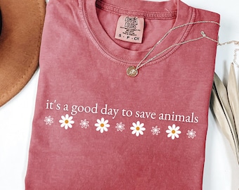 Könnte beinhalten: Staubrosafarbenes T-Shirt mit dem weißen Schriftzug "it's a good day to save animals" und Gänseblümchen-Grafiken. Eine silberne Halskette mit Anhänger und ein brauner Hut sind ebenfalls zu sehen.