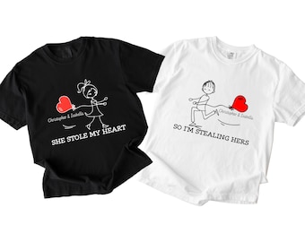 Puede incluir: Dos camisetas a juego, una negra y otra blanca, con un diseño caprichoso. La camiseta negra dice "She Stole My Heart" y la camiseta blanca dice "So I'm Stealing Hers". Cada camiseta presenta una figura de palo sosteniendo un globo de corazón rojo.