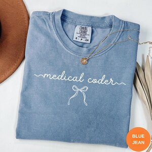 Könnte beinhalten: Ein blaues kurzärmeliges T-Shirt mit Rundhalsausschnitt und einem Comfort Colors-Etikett. Auf der Vorderseite befindet sich der weiße Schriftzug "medical coder" in Schreibschrift, ergänzt durch eine einfache weiße Liniengrafik einer Schleife.