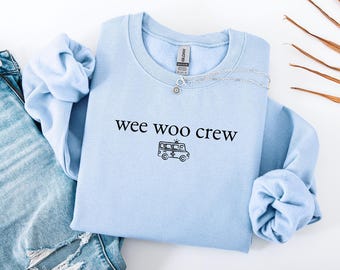 May include: A light blue crewneck sweatshirt with the text "wee woo crew" and an ambulance graphic in black. A silver necklace with a small pendant is visible. The sweatshirt is folded on top of a pair of blue jeans.