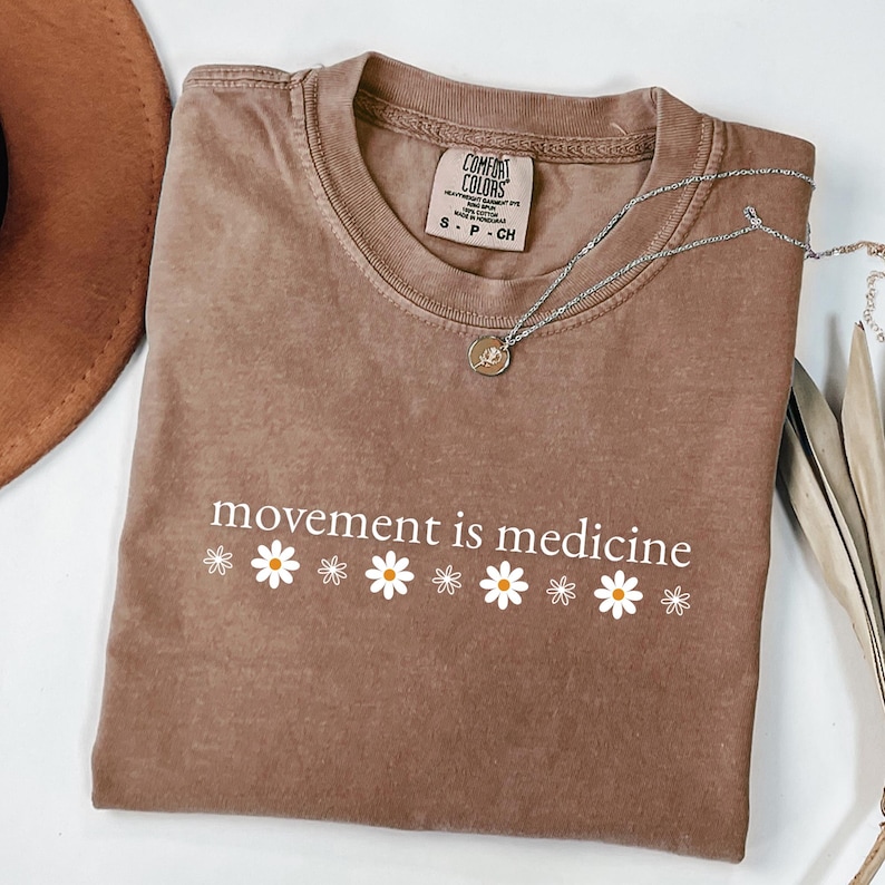 May include: A brown Comfort Colors t-shirt with the phrase "movement is medicine" in white script, accompanied by white daisy graphics. A silver necklace with a pendant is draped across the shirt.