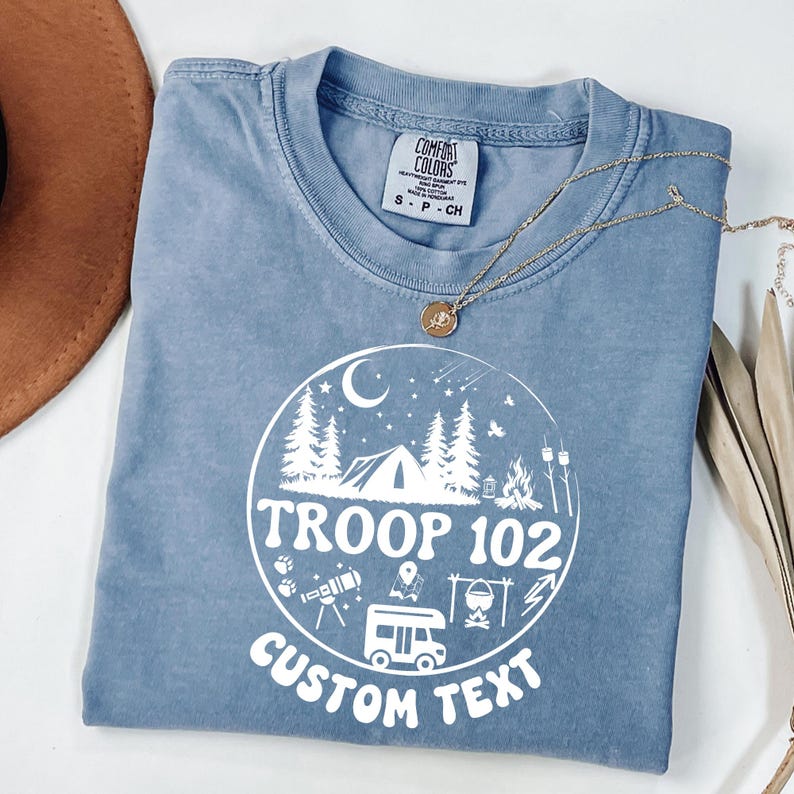 以下が含まれることがあります： 「TROOP 102」と「CUSTOM TEXT」の文字とキャンプシーンが描かれた白いグラフィックが特徴の、青色のComfort Colors Tシャツ。 金色のネックレスがシャツの上にかけられています。 シャツのサイズはS-CHです。