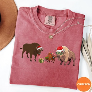 May include: A crimson short-sleeve t-shirt with a graphic print of two bison and a smaller bison calf. One bison wears a Santa hat and red bow, the other wears a Santa hat and green glasses. The calf is wrapped in colorful Christmas lights.