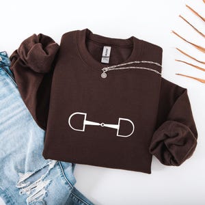 May include: A dark brown sweatshirt with a white horse bit graphic. A silver necklace with a circular pendant is draped across the sweatshirt. The sweatshirt is folded, with a pair of blue jeans visible in the background.
