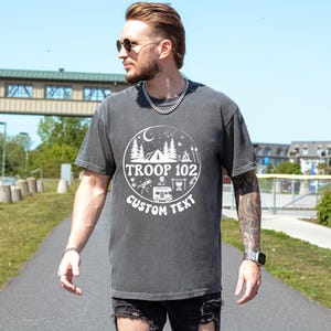 以下が含まれることがあります： 白い円形グラフィックが特徴の濃いグレーのTシャツ。テント、木、そして「TROOP 102」と「CUSTOM TEXT」の文字が描かれています。 このシャツは半袖で、クルーネックです。