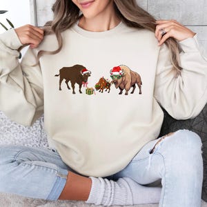 May include: A cream-colored crewneck sweatshirt featuring a graphic print of three bison. The left bison wears a Santa hat and a red bow, the center bison is wrapped in colorful Christmas lights, and the right bison wears a Santa hat and green glasses.