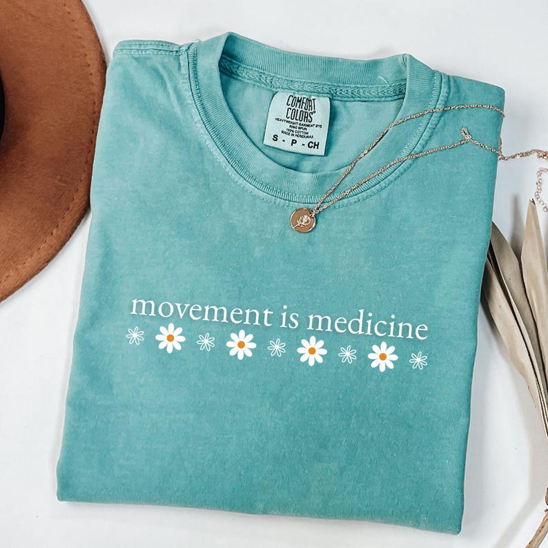 May include: A teal Comfort Colors t-shirt with the phrase "movement is medicine" in white, with daisy accents. A gold necklace with a pendant is draped over the shirt. A brown hat is visible in the upper left corner.