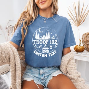 以下が含まれることがあります： 白い円形のグラフィックにキャンプ要素と「TROOP 102 CUSTOM TEXT」の文字が入った、青い半袖Tシャツ。デザインには、木、テント、キャンプファイヤー、キャンピングカーが含まれています。