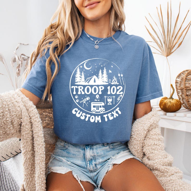 以下が含まれることがあります： 白い円形のグラフィックにキャンプ要素と「TROOP 102 CUSTOM TEXT」の文字が入った、青い半袖Tシャツ。デザインには、木、テント、キャンプファイヤー、キャンピングカーが含まれています。