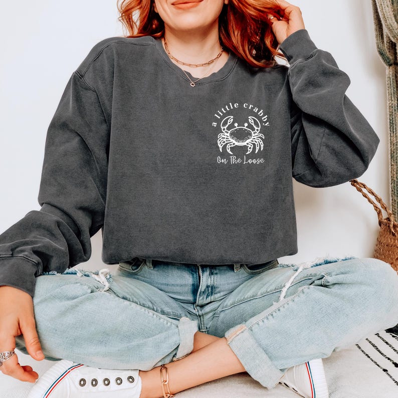 May include: Charcoal gray sweatshirt with a white crab graphic and the text "a little crabby On The Loose." The long-sleeved, crew neck sweatshirt is made of a soft material. The sweatshirt is paired with light blue jeans and white sneakers.