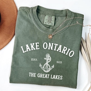 May include: An olive green t-shirt with the words "Lake Ontario" in white, along with an anchor graphic and the text "The Great Lakes." A gold necklace with a pendant rests on the shirt. A brown hat is visible in the upper left corner.