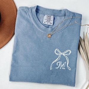 Peut inclure: Un t-shirt bleu Comfort Colors avec un nœud blanc et la lettre "M" imprimée. Un collier doré avec un pendentif repose sur le t-shirt. Un chapeau marron est visible.