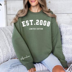 May include: An olive green sweatshirt with the text "EST. 2008 LIMITED EDITION" in white block letters. The name "Amelia" is written in cursive on the left sleeve. The sweatshirt is a casual style.