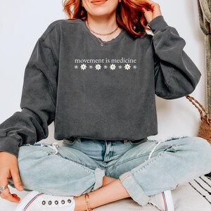May include: Dark gray sweatshirt with the text "movement is medicine" in white, with daisy accents. The sweatshirt has a relaxed fit and long sleeves. The model is wearing light blue jeans and white sneakers.