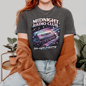May include: Dark gray t-shirt featuring a retro design with the text "MIDNIGHT RADIO CLUB" and a cassette tape graphic. Below the graphic is the text "late night frequency". The design has a vibrant color scheme with a vintage aesthetic.