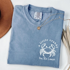 May include: A blue Comfort Colors t-shirt with a white crab graphic and the text "a little crabby On The Loose." A gold necklace with a pendant is draped across the shirt. A brown hat is visible in the upper left corner.