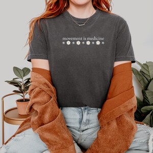 May include: Dark gray t-shirt with the text "movement is medicine" above a row of white daisy graphics. The shirt is paired with a rust-colored jacket and blue jeans. The overall aesthetic is casual and comfortable.