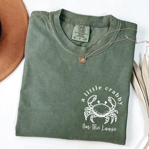 May include: Olive green Comfort Colors t-shirt with the text "a little crabby On The Loose" and a crab graphic. A gold necklace with a pendant is draped across the shirt. A brown hat is visible in the upper left corner.