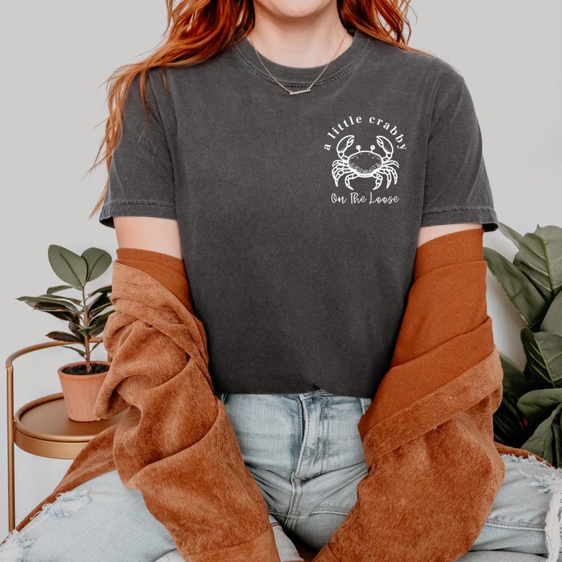 May include: Dark gray t-shirt with a white crab graphic and the text "a little crabby On The Loose." The shirt is made of a soft material and has a relaxed fit. The shirt is a casual wear item.