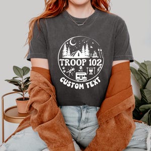 以下が含まれることがあります： テント、木、および「TROOP 102」のテキストが特徴の白い円形グラフィックが付いた濃い灰色のTシャツ。「CUSTOM TEXT」という言葉が円の下に表示されています。 このシャツはカジュアルなスタイルです。