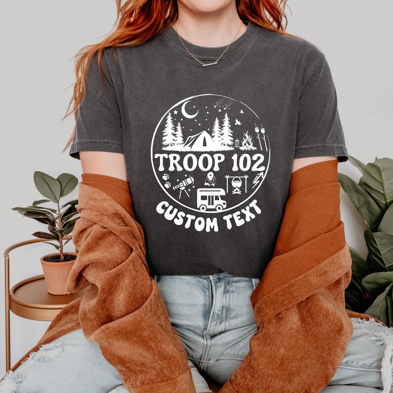 以下が含まれることがあります： テント、木、および「TROOP 102」のテキストが特徴の白い円形グラフィックが付いた濃い灰色のTシャツ。「CUSTOM TEXT」という言葉が円の下に表示されています。 このシャツはカジュアルなスタイルです。