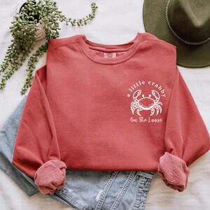 May include: A coral-colored sweatshirt with the phrase "a little crabby on the loose" and a white crab graphic. The long-sleeved top is folded over denim shorts, with a green hat and plant in the background.