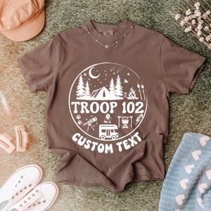 以下が含まれることがあります： テント、木、テキスト「TROOP 102 CUSTOM TEXT」が描かれた白い円形グラフィックが特徴の茶色のTシャツ。デザインには、望遠鏡、キャンプファイヤー、キャンピングカーが含まれています。シャツは帽子、靴、毛布に囲まれています。
