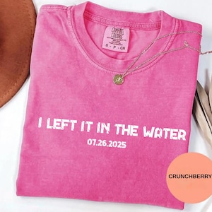 Puede incluir: Una camiseta rosa brillante con el texto blanco "I LEFT IT IN THE WATER" y la fecha "07.26.2025". La camiseta tiene cuello redondo y mangas cortas. Un collar dorado con un colgante está sobre la camiseta. La palabra "CRUNCHBERRY" está en un círculo.