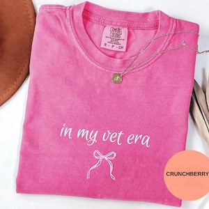 May include: A bright pink Comfort Colors t-shirt with the text "in my vet era" and a bow graphic. A gold necklace with a pendant is draped across the shirt. The shirt is a size small.