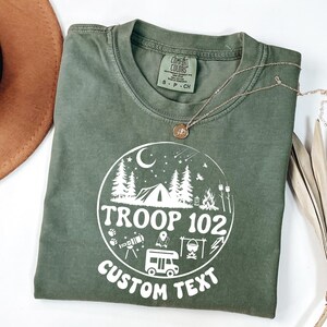 以下が含まれることがあります： オリーブグリーンのTシャツ。テント、木、そして「TROOP 102 CUSTOM TEXT」の文字が描かれた円形のグラフィックが特徴です。ペンダント付きのゴールドネックレスがシャツの上にかけられています。シャツはComfort Colors素材です。