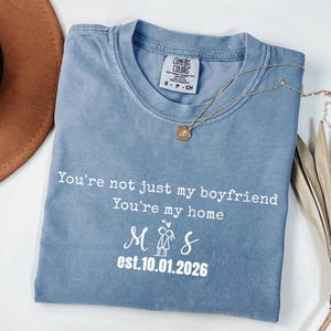 Op de afbeelding: Een blauw Comfort Colors t-shirt met witte tekst: "You're not just my boyfriend, You're my home". Het shirt toont ook de initialen "M & S" en de datum "est.10.01.2026". Een gouden ketting met hanger ligt op het shirt.