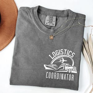 Puede incluir: Una camiseta gris Comfort Colors con un gráfico blanco que dice "Logistics Coordinator". El gráfico incluye un avión, un barco y un camión. Un collar plateado con un pequeño colgante está sobre la camiseta.