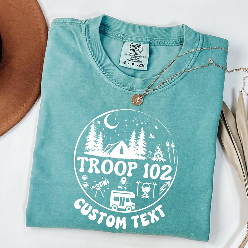 以下が含まれることがあります： ターコイズブルーのComfort Colors Tシャツ。白いキャンプをテーマにしたグラフィックが特徴で、テント、木、キャンプファイヤー、テキスト「TROOP 102 CUSTOM TEXT」が含まれています。 金色のペンダント付きネックレスがシャツの上にかけられています。