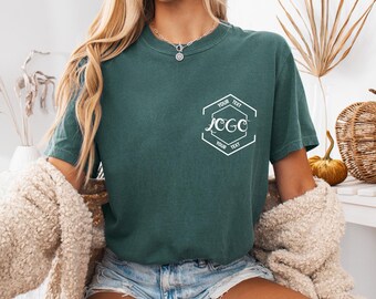 以下が含まれることがあります： 左胸に白いグラフィックがプリントされたダークグリーンの半袖Tシャツ。グラフィックは六角形の輪郭で構成されており、上下に「YOUR TEXT」という文字、中央に筆記体で「LOGO」という文字がデザインされています。