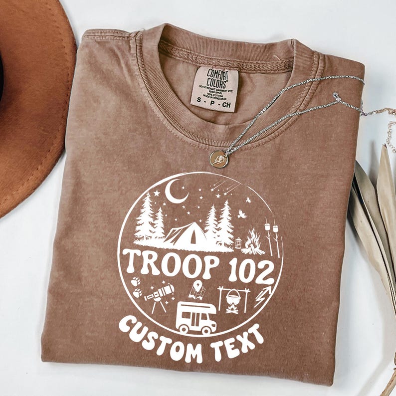 以下が含まれることがあります： 白のキャンプをテーマにしたグラフィックが特徴の、茶色のComfort Colors Tシャツ。グラフィックにはテント、木、キャンプファイヤー、テキスト「TROOP 102 CUSTOM TEXT」が含まれています。ペンダント付きのシルバーネックレスも表示されています。