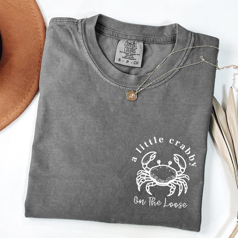 May include: A gray Comfort Colors t-shirt with the phrase "a little crabby On The Loose" and a crab graphic. A gold pendant necklace and a brown hat are also visible.