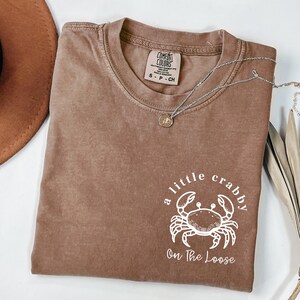 May include: A brown t-shirt with the text "a little crabby On The Loose" and a white crab graphic. The shirt has a crew neck and short sleeves. A silver necklace with a small pendant is on the shirt.