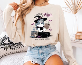 May include: Cream-colored long-sleeve shirt featuring a graphic of a witch reading a book, with the text "Just a Witch Who Loves Books." The witch is depicted with blue hair, a black hat, and glasses. Purple bats and flames are also in the design.