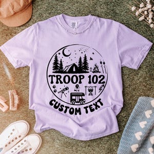 以下が含まれることがあります： テント、木、そして「TROOP 102 CUSTOM TEXT」の文字が描かれた円形グラフィックが特徴の薄紫色のTシャツ。キャンプファイヤーやキャンピングカーなど、キャンプをテーマにしたイラストがデザインされています。シャツはクルーネックです。