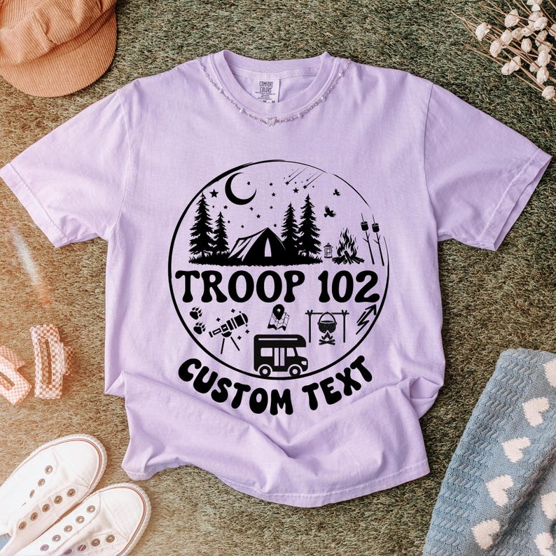 以下が含まれることがあります： テント、木、そして「TROOP 102 CUSTOM TEXT」の文字が描かれた円形グラフィックが特徴の薄紫色のTシャツ。キャンプファイヤーやキャンピングカーなど、キャンプをテーマにしたイラストがデザインされています。シャツはクルーネックです。