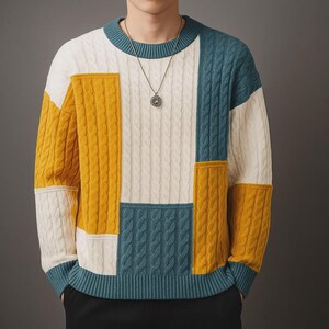 Op de afbeelding: Een colorblock trui in een kabelgebreid ontwerp. De trui heeft panelen in wit, mosterdgeel en blauwgroen. De halslijn en manchetten zijn blauwgroen. Een zilveren hanger hangt aan een ketting.