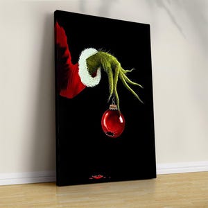 Könnte beinhalten: Leinwanddruck mit einer grünen, krallenartigen Hand mit weiß getrimmtem rotem Ärmel, die eine rote Weihnachtskugel vor schwarzem Hintergrund hält. Das Kunstwerk lehnt an einer hellbraunen Holzoberfläche.