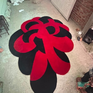 May include: A large, irregularly shaped area rug featuring a bold, abstract pattern of interlocking organic forms in vibrant red and deep black. The rug has a soft, tufted texture and a curved, flowing silhouette that covers a significant floor area.