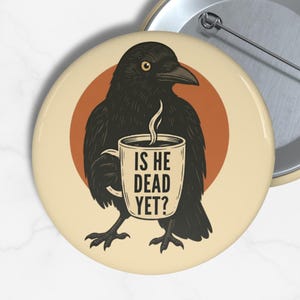 May include: A round button with a black crow illustration holding a coffee mug. The mug has the text "IS HE DEAD YET?". The crow is set against a brown circle on a cream background. A second button is visible.